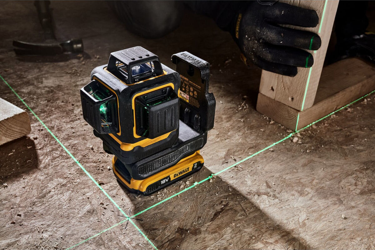 Laser krzyżowy DEWALT DCLE34031D1-QW, optyczne, elementy, IP54, pył, wilgoć, laser stoi na płycie OSB, w tle dłoń trzyma kawałek drewna