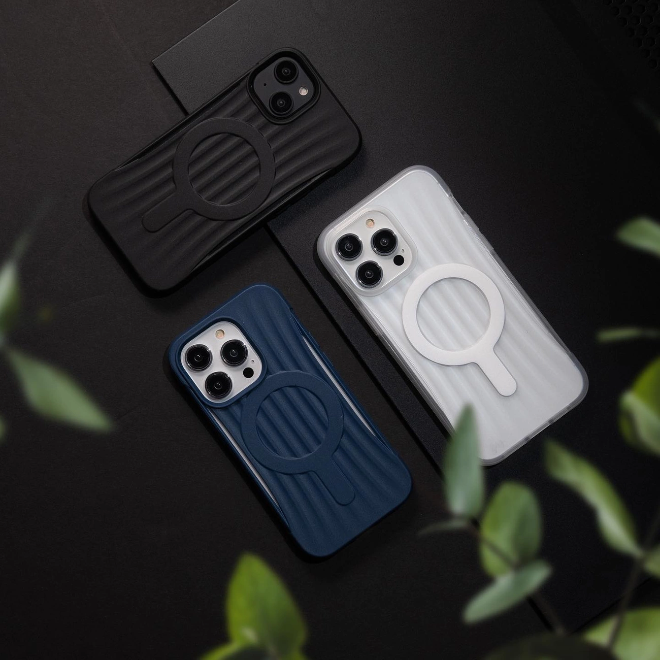Na obrazie widoczne są trzy etui na telefon w kolorach czarnym, niebieskim i białym, każde z okrągłym wzorem na tylnej części. Etui leżą na ciemnym tle, a w dolnym rogu widoczne są rozmyte zielone liście.