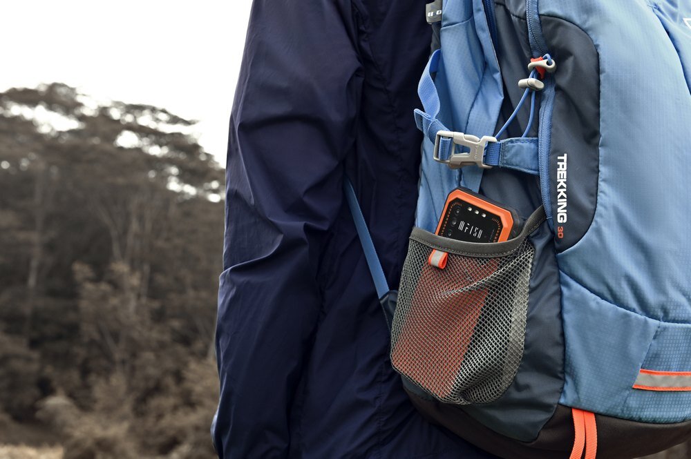 Osoba w niebieskiej kurtce nosi niebieski plecak z napisem \'TREKKING\', w którego siatkowej kieszeni znajduje się pomarańczowy smartfon. W tle widoczne są drzewa.