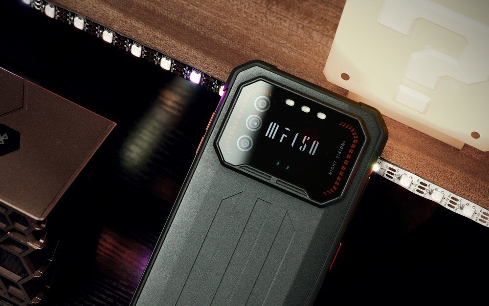Na obrazie widoczny jest smartfon z czarną obudową i wyświetlaczem z napisem \'IIIF150\'. W tle znajdują się elementy dekoracyjne i oświetlenie LED.