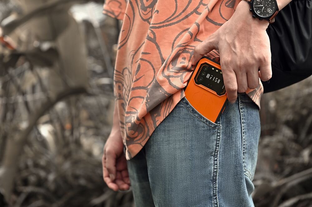 Osoba wkłada pomarańczowy smartfon do kieszeni spodni dżinsowych. Na nadgarstku ma zegarek, a koszulka ma wzór w kwiaty.