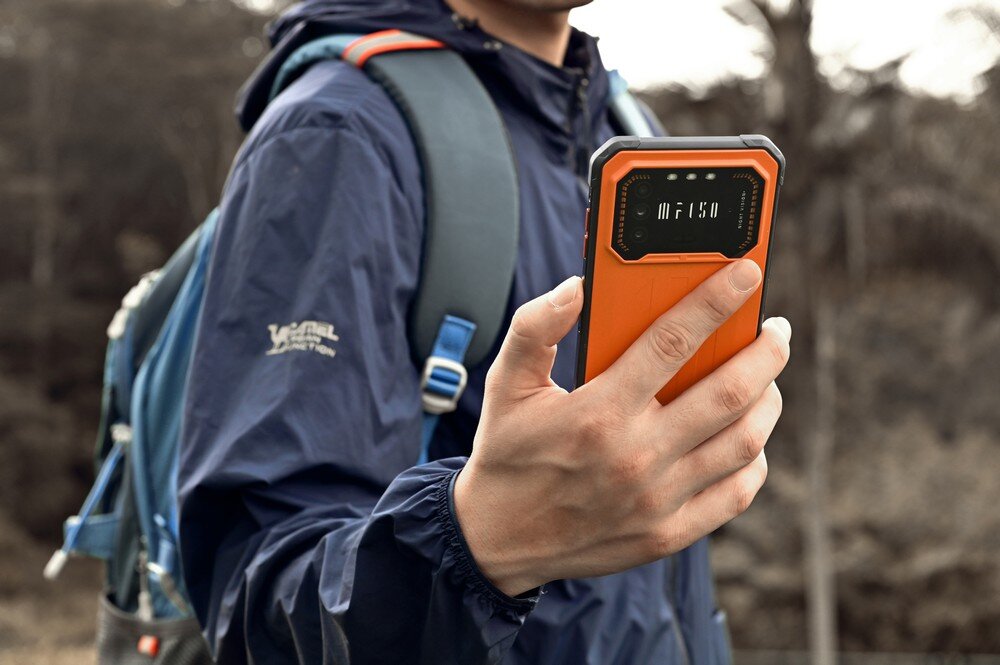 Osoba trzyma pomarańczowy smartfon z widocznym napisem \'IIIF150\' na tylnej obudowie. W tle znajduje się rozmyty krajobraz z drzewami.