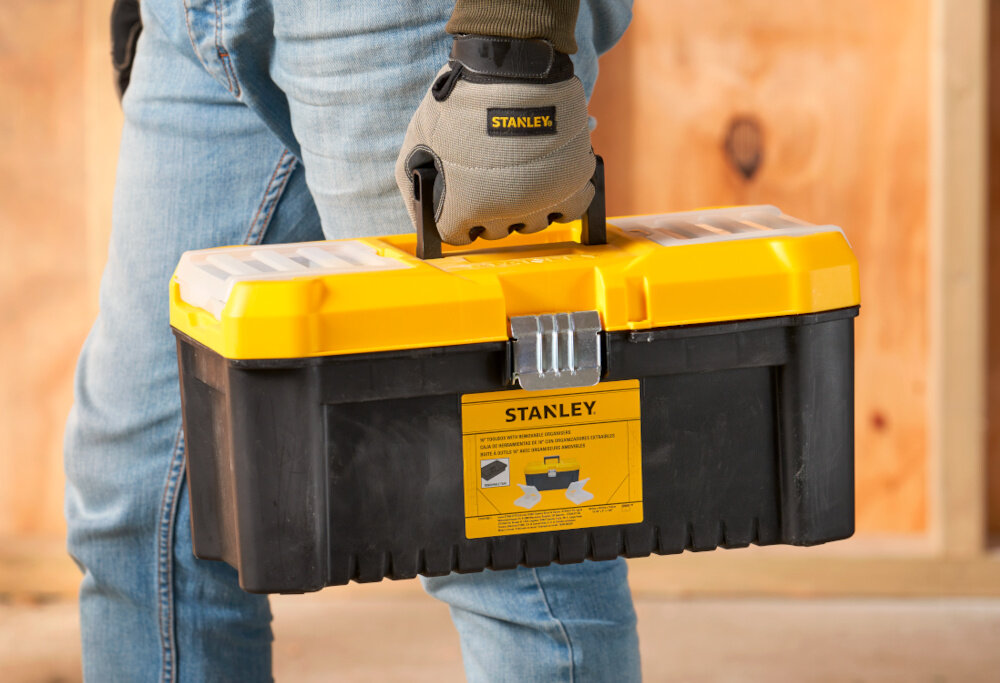 Skrzynka narzędziowa STANLEY Essential 16‘’ STST75785-1 osoba niesie w dłoni skrzynkę w tle drewniana ściana duża rękojeść z wysokiej jakości tworzywa sztucznego stabilny chwyt komfort przenoszenia ergonomiczny kształt pewnie leży w dłoni nie ślizga się w czasie transportu