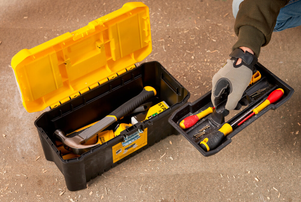 Skrzynka narzędziowa STANLEY Essential 16‘’ STST75785-1 skrzynia na drewnianym blacie wypełniona narzędziami obok osoba wyciąga ze skrzyni tackę wyjmowana tacka łatwy dostęp ułatwia utrzymanie porządku