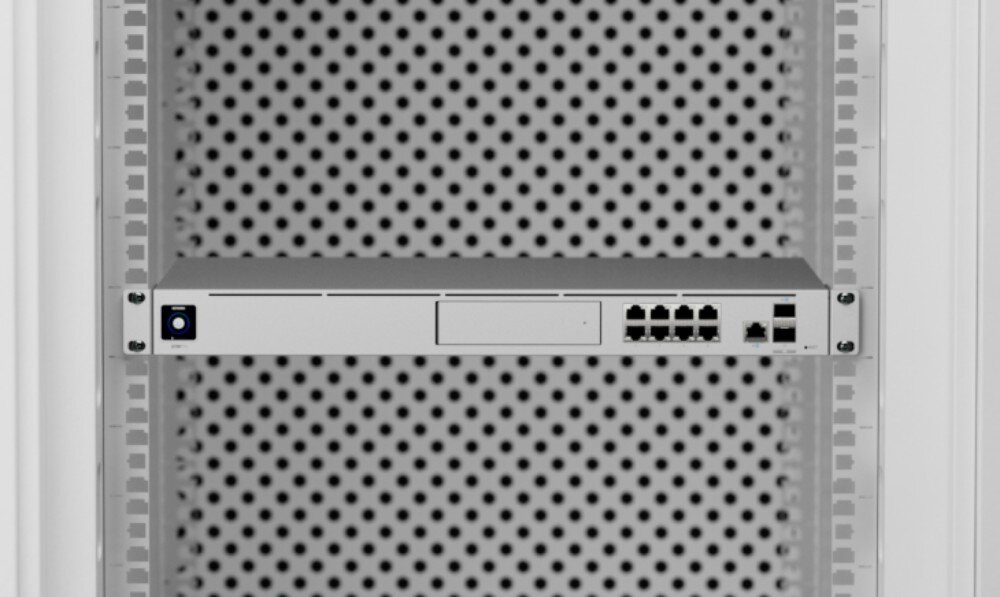Router UBIQUITI UDM-SE-EU Front srebrnego urządzenia sieciowego zamontowanego w szafie rack na tle ciemnej perforowanej blachy, ukazujący porty Ethernet i mały ekran po lewej stronie