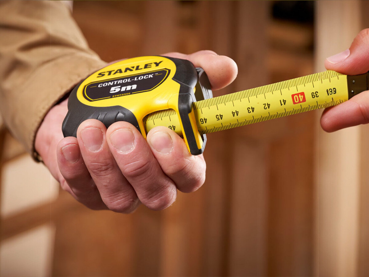 Osoba trzyma miarę zwijaną z widocznym logo \'STANLEY\' i napisem \'CONTROL-LOCK 5m\'. Taśma miary jest wysunięta i pokazuje oznaczenia długości w centymetrach.