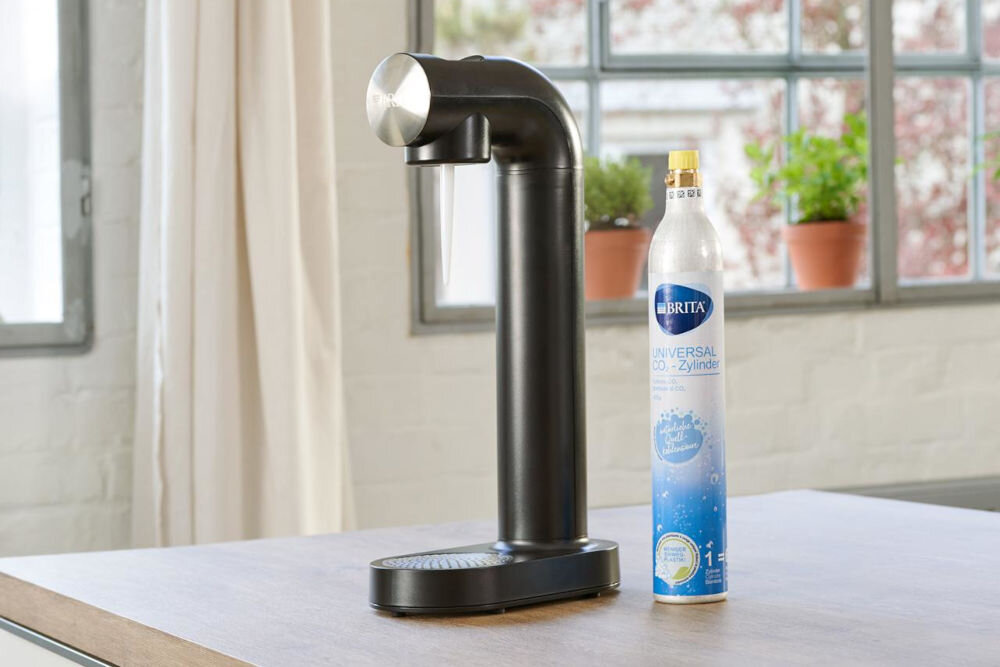 Nabój BRITA Sodaone CO2 1043744 na jasnym blacie stoi czarny saturator Brita SodaOne, a obok niego srebrny cylinder z dwutlenkiem węgla. W tle widać okno z roślinami