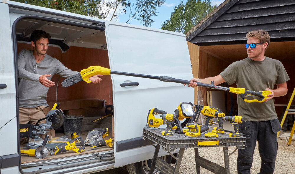 Piła akumulatorowa STANLEY Fatmax SFMCPS620M1 dwie osoby jedna osoba podaje drugiej osobie piłę w tle drewniana szopa do pielęgnacji drzew i krzewów zasilanie akumulatorowe wspiera codzienne prace porządkowe