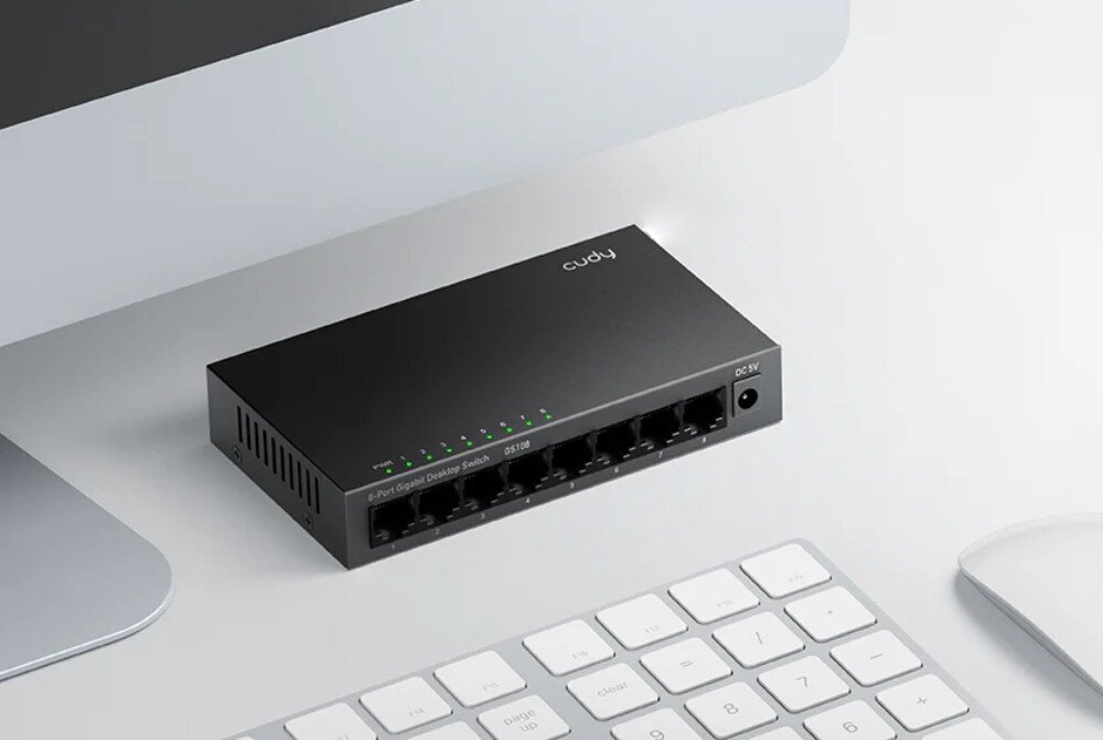 Switch CUDY GS108 Przełącznik ustawiony na biurku obok komputera i klawiatury. Podłączony zasilacz i zapalone zielone diody LED nad portami, wielkość, wymiary