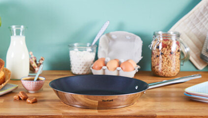 Na drewnianym blacie kuchennym znajduje się patelnia z metalowym uchwytem, a w tle widać różne produkty spożywcze, takie jak jajka, mleko i płatki zbożowe w słoiku. Na patelni widoczne jest logo \'Tefal\'.