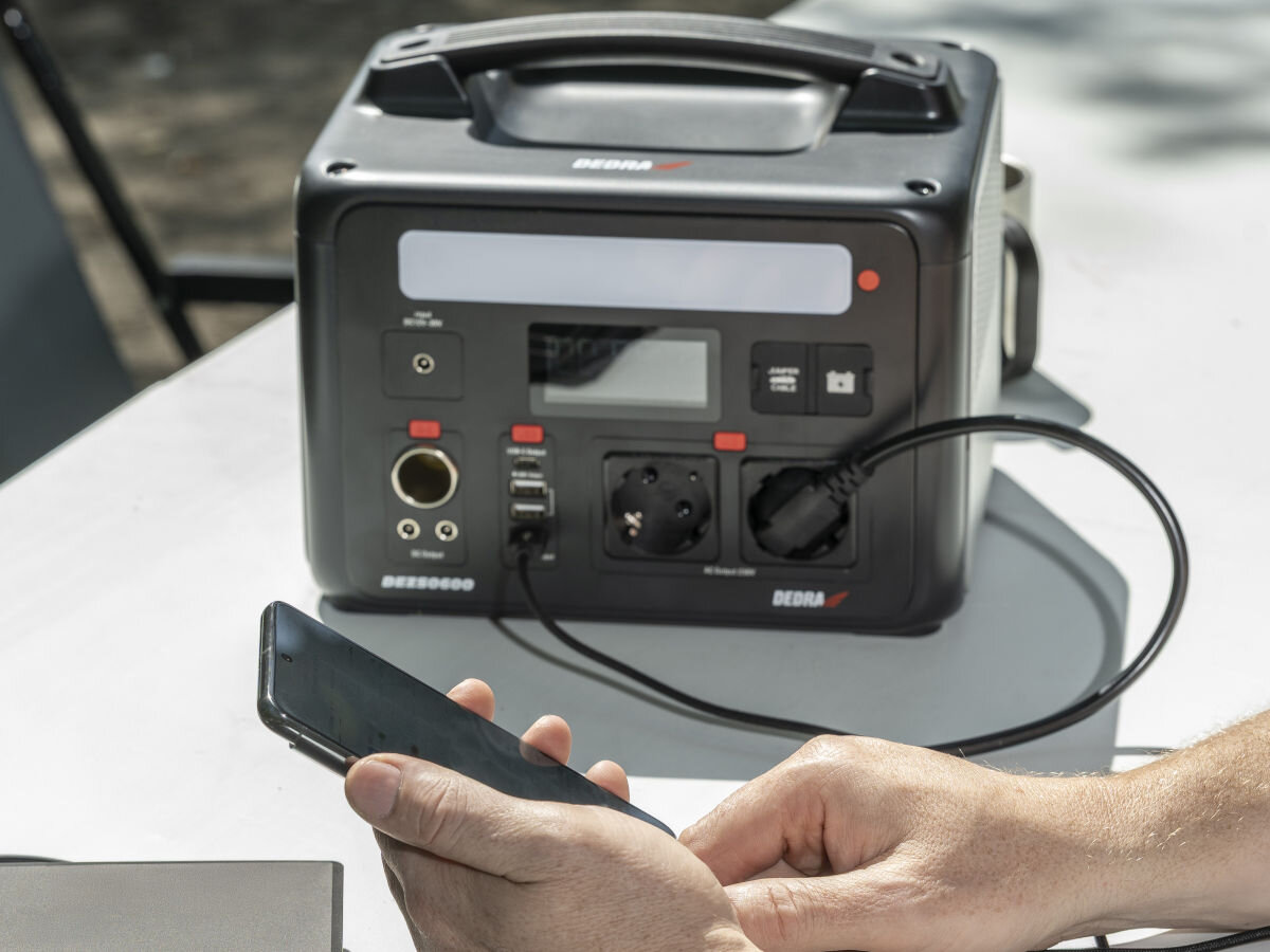Stacja zasilania DEDRA DEZS0600 600W Bank energii porty gniazda wejścia oświetlenie, stacja stoi na stole składanym, w tle osoba podpina smartfona do stacji