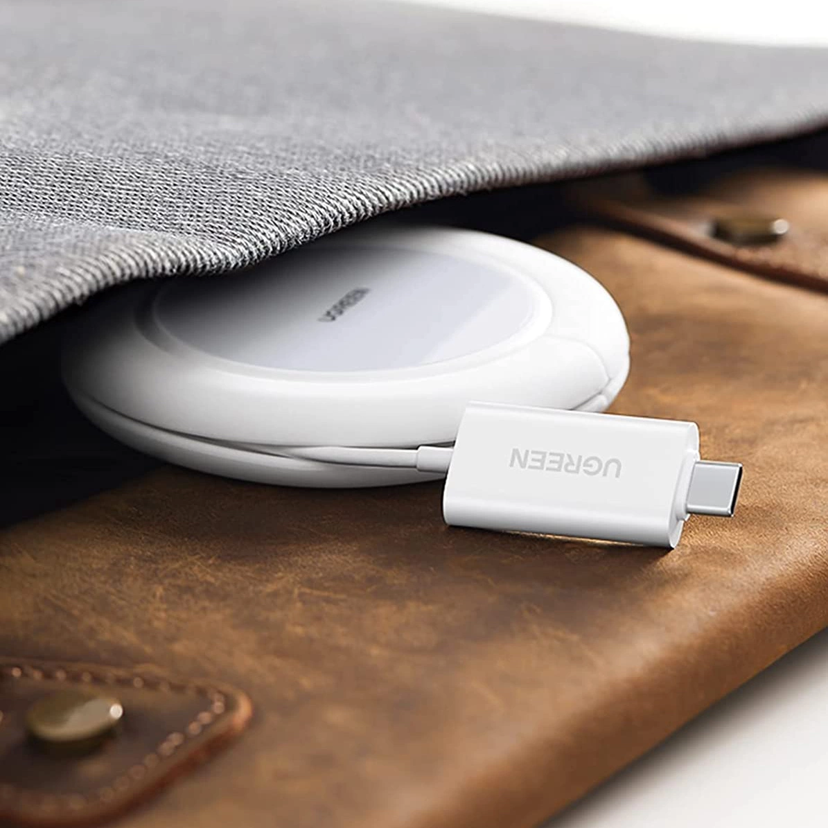 Biała ładowarka bezprzewodowa z kablem USB-C marki UGREEN jest częściowo schowana pod szarym materiałem na brązowej powierzchni. Na kablu widoczne jest logo \'UGREEN\'.