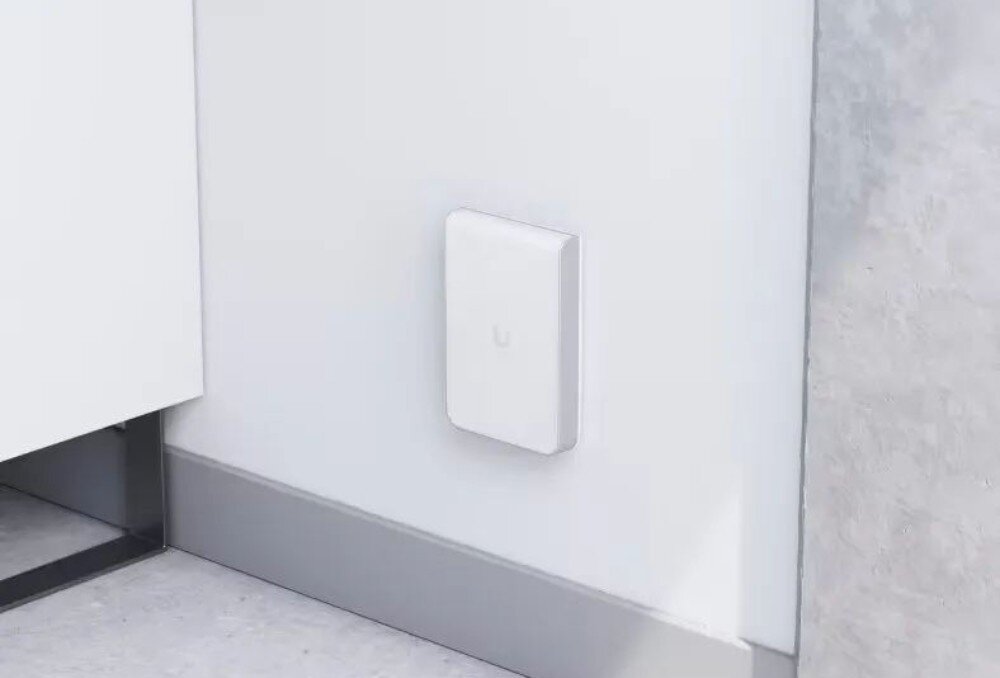 Punkt dostępu UBIQUITI U6-IW Biały prostokątny punkt dostępowy zamontowany na ścianie tuż nad listwą podłogową w jasnym pomieszczeniu