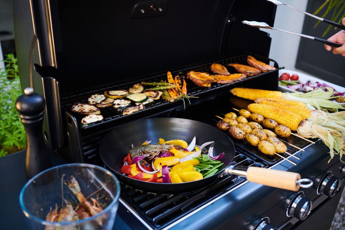 Grill gazowy CAMPINGAZ Select LX PLUS Seria 4 12 kW 46 x 72 cm, żeliwo, emaliowane, wymiary, temperatura, w tle ogród, zbliżenie na ruszty grilla na którym jest różnego rodzaju potraw