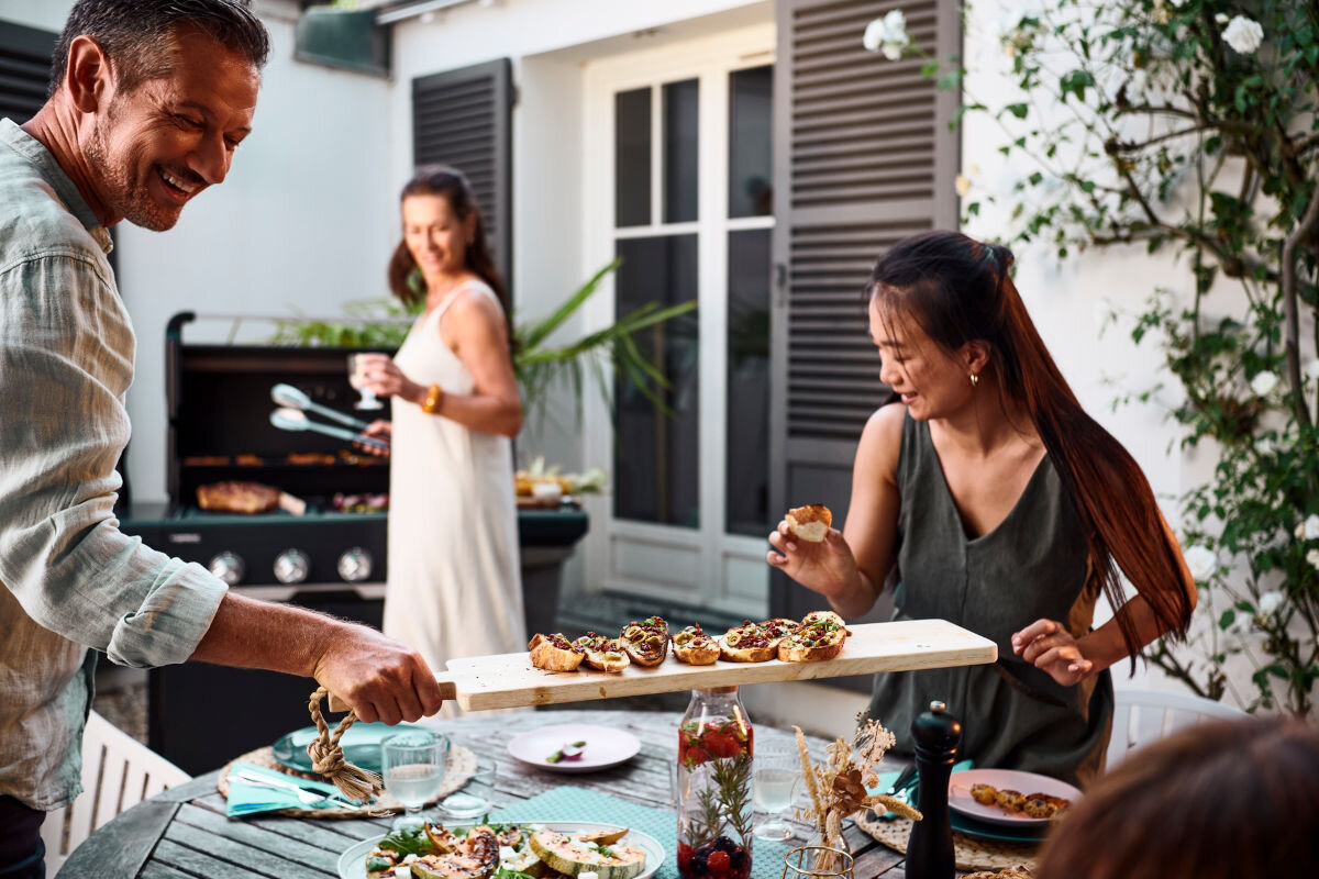 Grill gazowy CAMPINGAZ Select LX PLUS Seria 4 12 kW 46 x 72 cm, zaprojektowany, wysokość, ruszt, komfortowa, osoby siedzą przy stole, rozmawiają, w tle kobieta pracuje przy grillu