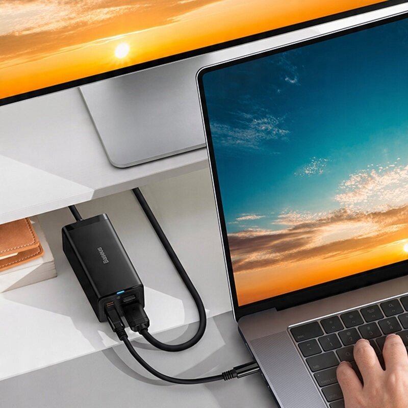 Na biurku znajduje się laptop z widocznym ekranem, obok którego leży czarna ładowarka z logo \'Baseus\'. Ładowarka jest podłączona do laptopa oraz do gniazdka za pomocą kabli.