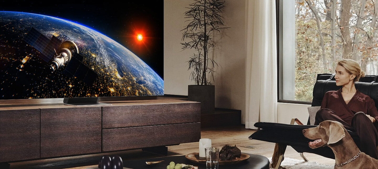 Telewizor Samsung Neo Qled 8K Excellence Line  stojące na szafce obok kobiety z psem siedzącej na kanapie. QN700BTXXH