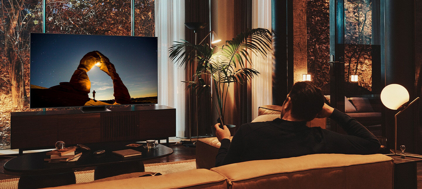 Telewizor Samsung Neo QLED 8K Excellence Line QN700B na szafce naprzeciwko mężczyzny siedzącego na kanapie. QN700BTXXH