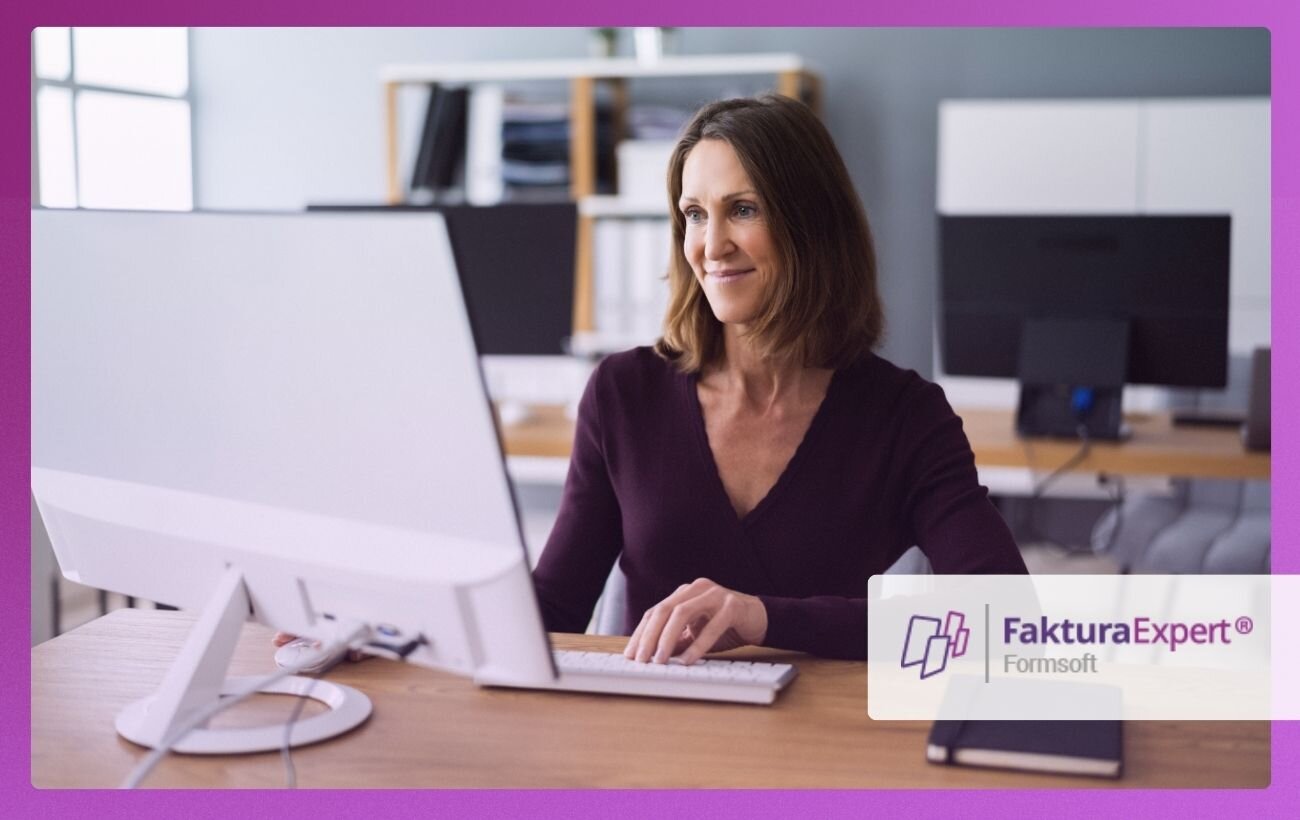 uśmiechnięta kobieta w&nbsp;średnim wieku i&nbsp;krótkich włosach pracuje w&nbsp;biurze; patrzy na ekran komputera i&nbsp;pisze na klawiaturze; w&nbsp;prawym dolnym rogu logo Faktura Expert.