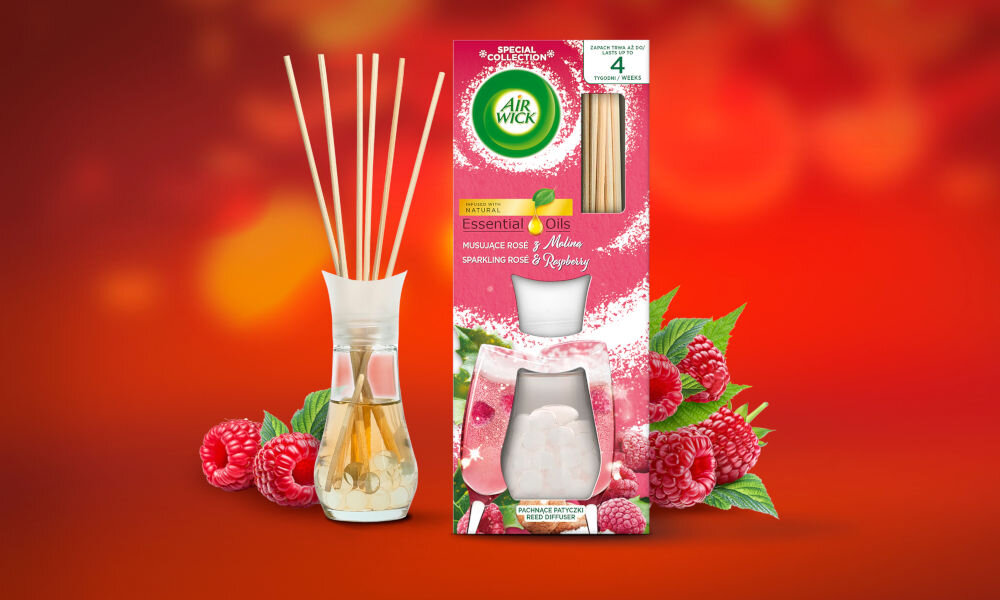 Na obrazie widoczne są patyczki zapachowe w szklanym flakonie oraz opakowanie produktu Air Wick Essential Oils z grafiką malin i róż. Na opakowaniu znajduje się tekst: \'Musujące Róże & Maliny Sparkling Rose & Raspberry\'.
