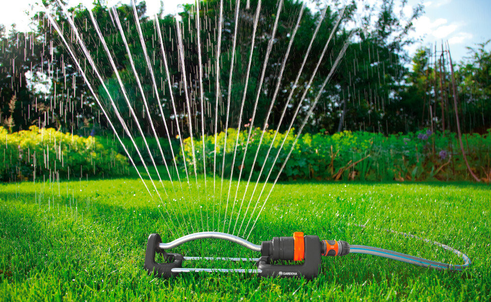Zraszacz GARDENA Aqua S 18700-20 trawnik zraszacz na trawniku w tle drzewa krzewy kwiaty zraszacz wahadłowy do równomiernego nawadniania trawników i ogrodów o powierzchni od 90 do 220 m² eliminuje potrzebę ręcznego podlewania oszczędność czasu i energii