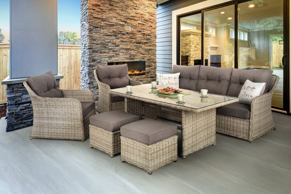 Zestaw mebli ogrodowych złożony z sofy, dwóch foteli, dwóch puf i stołu, wykonany z plecionki w kolorze szarym. Na poduszkach widoczny jest tekst \'Home is where\'.