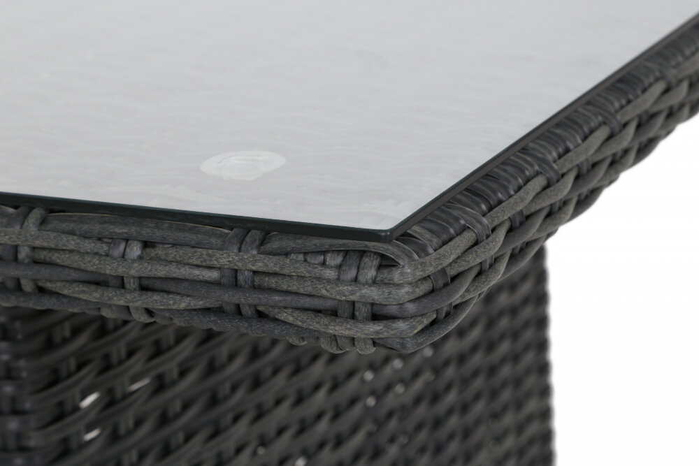 Zbliżenie na róg stołu ogrodowego z plecionki w ciemnym kolorze, z przezroczystym szklanym blatem.