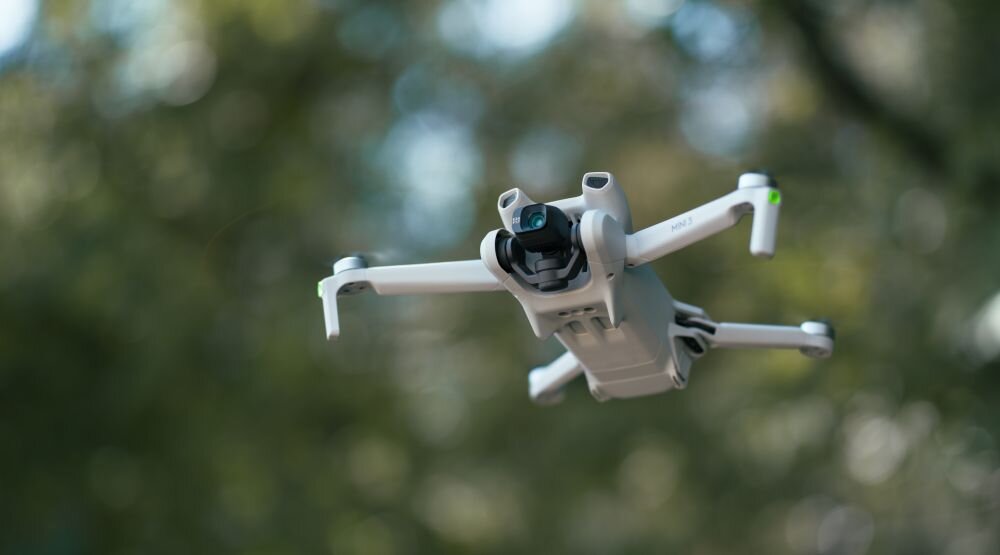 Dron DJI Mini 3 Szary dron uchwycony w locie od dołu, pokazujący podwozie oraz kamerę skierowaną przed siebie na tle rozmytego, zielonego lasu, czas lotu, transmisja obrazu, zasięg