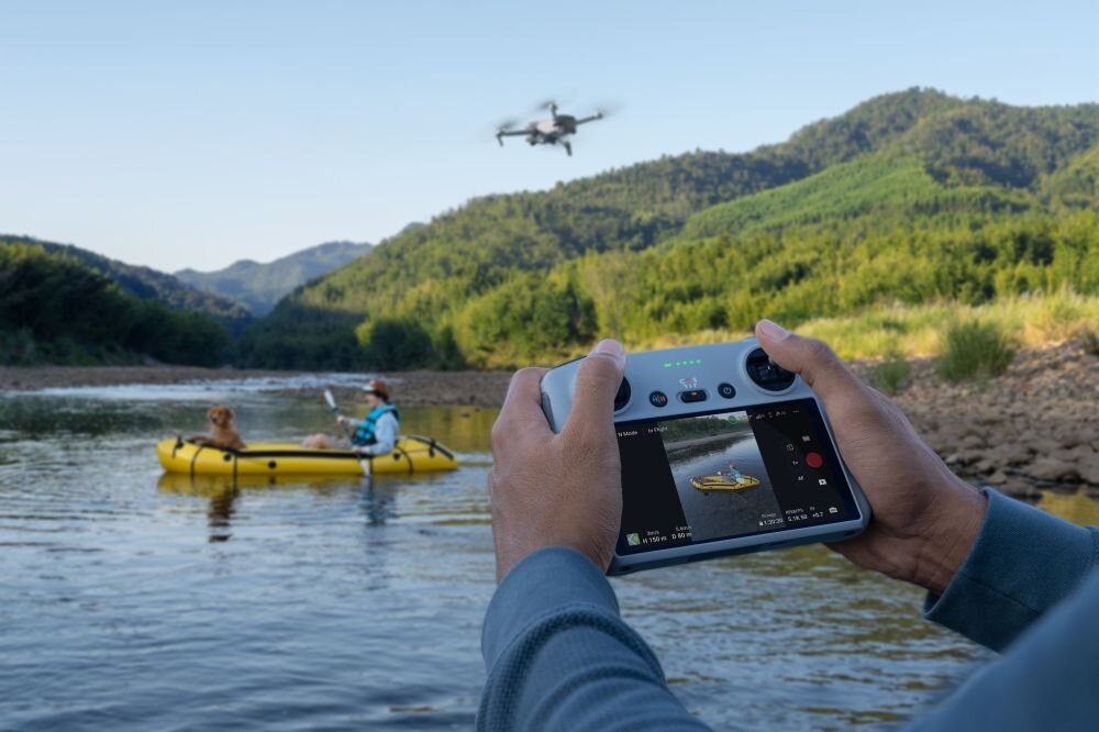Dron DJI Mini 3 Dłonie trzymają aparaturę sterującą z włączonym ekranem, na którym widać podgląd rzeki i żółtego pontonu, podczas gdy w oddali nad wodą leci mały dron, sterowanie, obsługa, asysty lotu