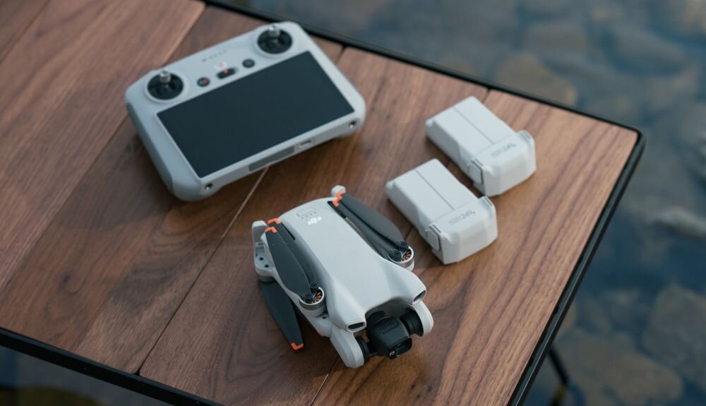 Dron DJI Mini 3 Zestaw akcesoriów składający się ze złożonego drona, kontrolera z ekranem oraz dwóch akumulatorów ułożonych na drewnianym stole blisko brzegu wody, aparatura sterująca, aplikacja, wyświetlacz