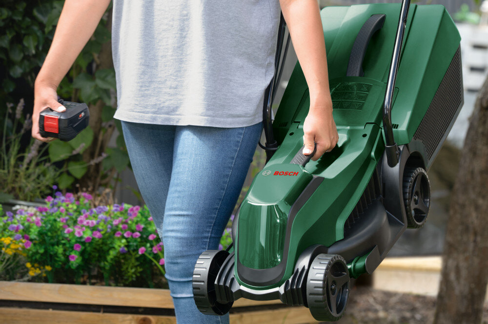 Kosiarka akumulatorowa BOSCH EasyMower 18V-32-200 06008B9D00 osoba niesie w dłoni kosiarkę w tle dom waga 8,1 kg zintegrowany uchwyt do przenoszenia lepsze efekty koszenia łatwie prowadzenie poziom hałasu na poziomie 88 dB intuicyjne sterowanie proste funkcje