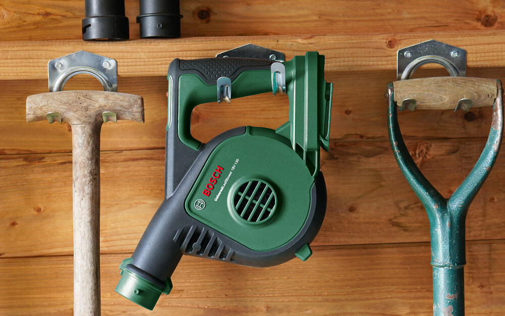 Dmuchawa do liści BOSCH UniversalLeafBlower szopa garaż drewniana ściana dmuchawa zawieszona na haku obok narzędzia ogrodowe waga około 1,6 kg komfortowy ergonomiczny uchwyt możliwość zawieszenia ułatwia przenoszenie i przechowywanie