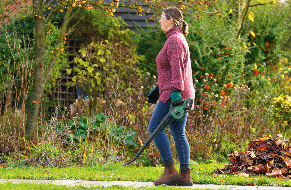 Dmuchawa do liści BOSCH UniversalLeafBlower ogród osoba idzie z dmuchawą w dłoni w tle dom krzewy drzewa wymiary 831 x 139 x 219 mm waga 1,3 kg bez akumulatora, 1,6 kg z akumulatorem łatwa w manewrowaniu i transportowaniu