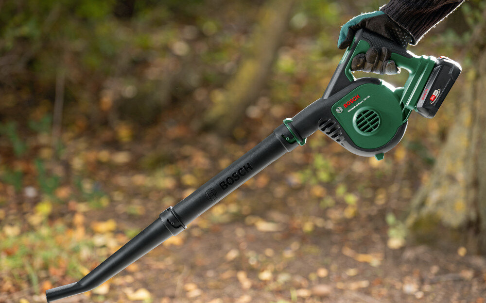 Dmuchawa do liści BOSCH UniversalLeafBlower drzewa liście osoba trzyma dmuchawę w dłoni generuje minimalną emisję hałasu i wibracji 0,0 m/s² niska emisja drgań chroni ręce i nadgarstki czas pracy na jednym ładowaniu około 18 minut