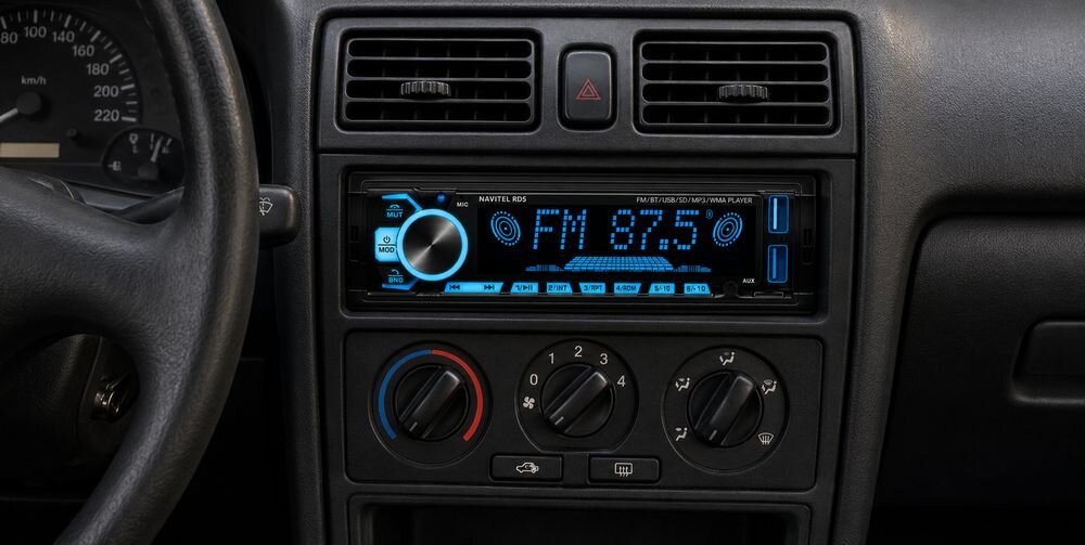 Obraz przedstawia radio samochodowe z wyświetlaczem pokazującym \'FM 87.5\' oraz smartfon z aplikacją muzyczną. Na środku znajduje się ikona symbolizująca połączenie Bluetooth.