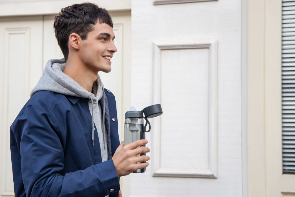 Butelka filtrująca PHILIPS GoZero AWP2731GRR/58 Profil uśmiechniętego mężczyzny w niebieskiej kurtce trzymającego otwartą butelkę filtrującą Philips na tle białej elewacji.