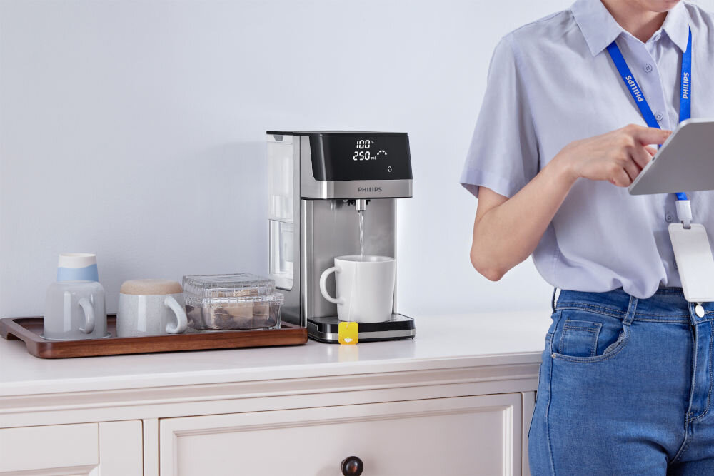 Dyspenser do wody PHILIPS ADD5910M/10 Kobieta przygotowuje herbatę, korzystając z dystrybutora wody Philips. Do białego kubka nalewana jest gorąca woda o temperaturze 100°C. Obok na tacy filiżanki i akcesoria biurowe.