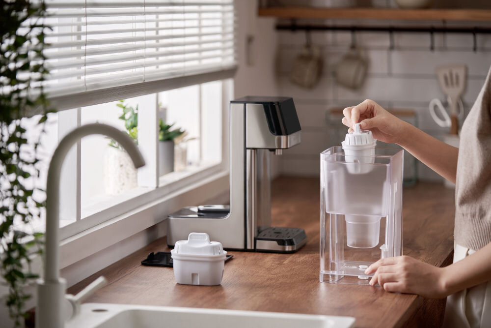 Dyspenser do wody PHILIPS ADD5910M/10 Prezentacja systemu filtracji: osoba wkłada biały wkład filtrujący do przezroczystego zbiornika, który zasila dystrybutor wody Philips. W tle widoczna jasna kuchnia.
