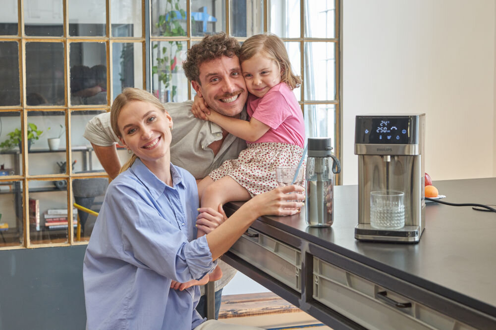 Dyspenser do wody PHILIPS ADD5910M/10 Uśmiechnięta rodzina z dzieckiem w kuchni przy wyspie, na której stoi dystrybutor wody Philips. Urządzenie nalewa czystą wodę do szklanki, promując zdrowe nawyki nawadniania.