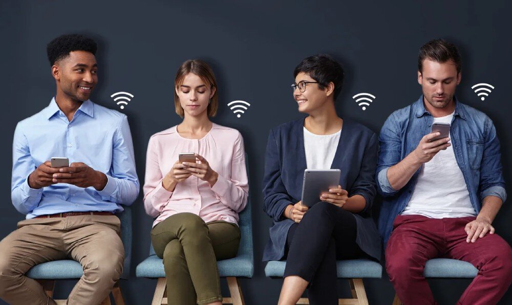 Cztery osoby siedzą na krzesłach, każda z urządzeniem mobilnym w ręku. Nad każdą osobą widoczna jest ikona sygnału Wi-Fi.