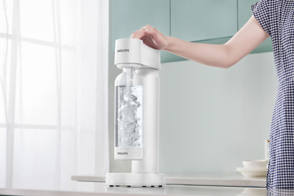 Saturator PHILIPS ADD4901WH/10 GoZero Zdjęcie prezentuje proces gazowania wody. Ręka użytkownika naciska górny przycisk białego saturatora Philips, a wewnątrz przezroczystej butelki widoczne jest intensywne powstawanie bąbelków dwutlenku węgla. Urządzenie stoi na jasnym blacie przy oknie.