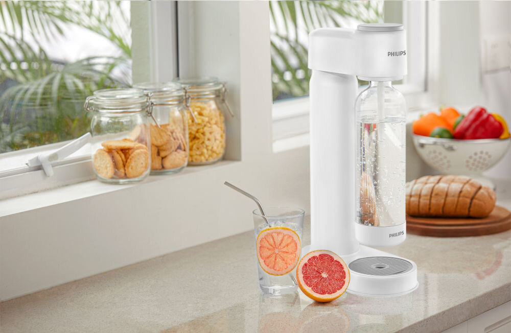 Saturator PHILIPS ADD4901WH/10 GoZero Biały saturator Philips z butelką pełną bąbelków stoi na jasnym blacie kuchennym przy oknie. Obok urządzenia znajduje się szklanka wody z plastrem grejpfruta i metalową słomką oraz połówka owocu. W tle widoczne są słoiki z przekąskami, świeży chleb i kolorowe papryki.