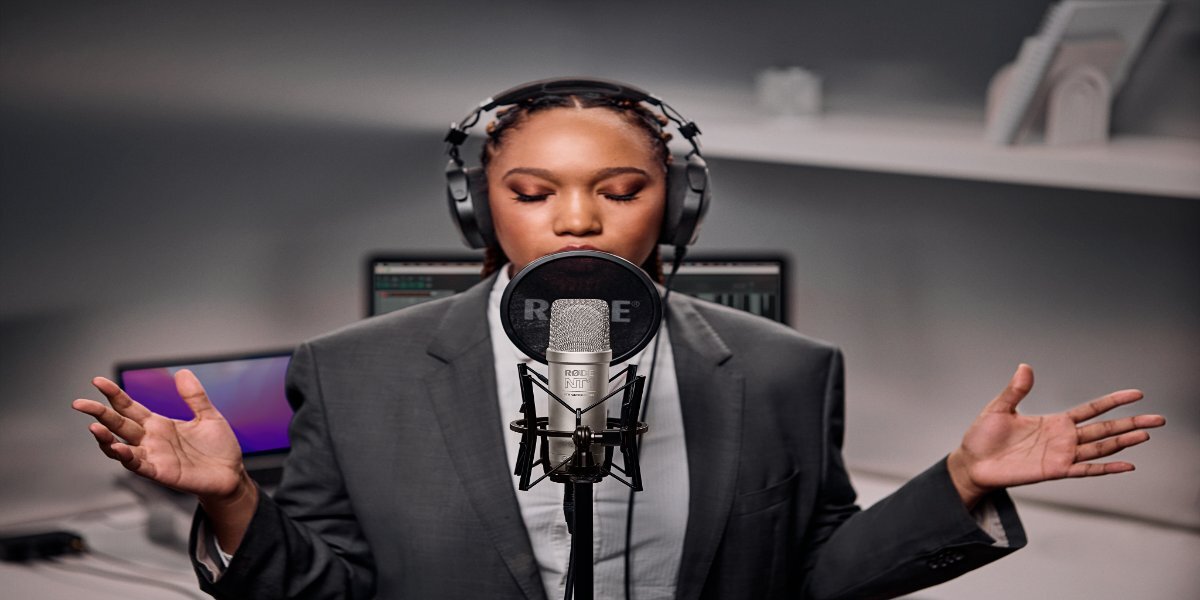 Osoba w słuchawkach stoi przed mikrofonem RODE NT1, trzymając ręce uniesione. W tle widoczne są monitory komputerowe.