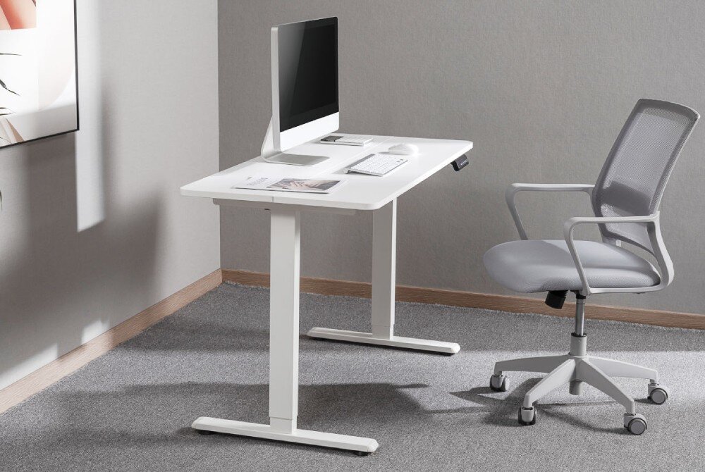 Stelaż biurka MOZOS EDF-PRO Biała metalowa rama biurka bez blatu ustawiona przy ścianie w jasnym pomieszczeniu obok kompletnie wyposażonego biurka z krzesłem i monitorem, opis