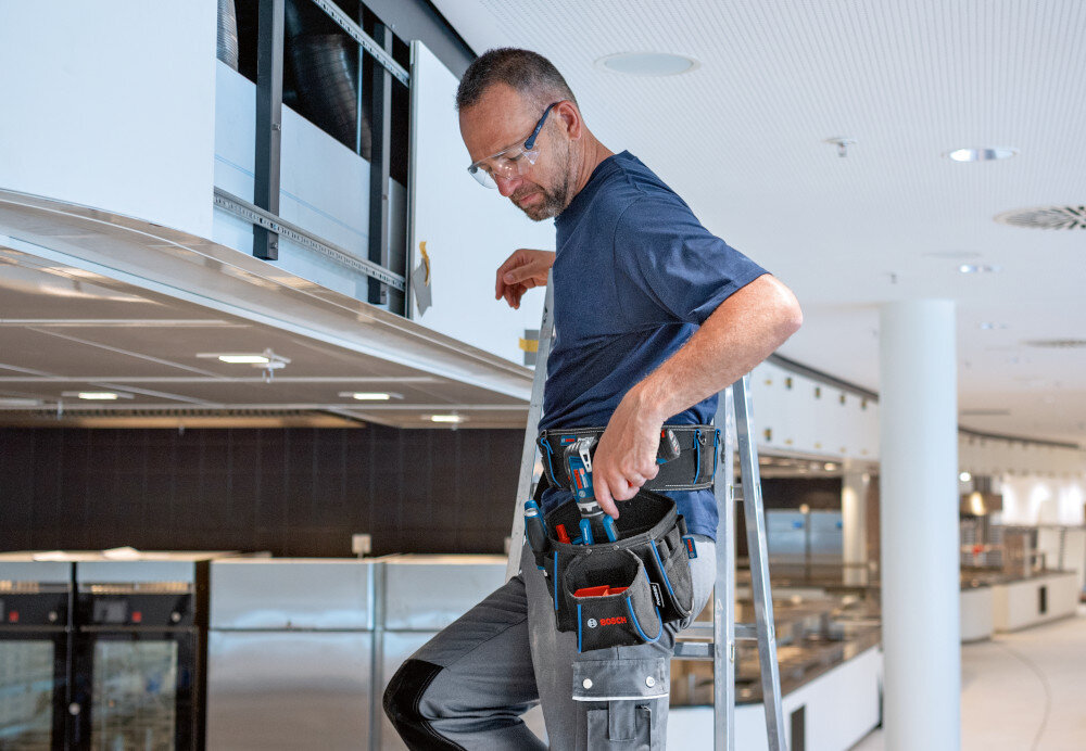 Torba narzędziowa BOSCH 1.600.A02.65T pomieszczenie osoba stoi na drabinie do pasa ma przymocowany pas i torbę wymiary 150 × 210 × 250 mm bardzo poręczna nie zajmuje wiele miejsca mobilność funkcjonalność