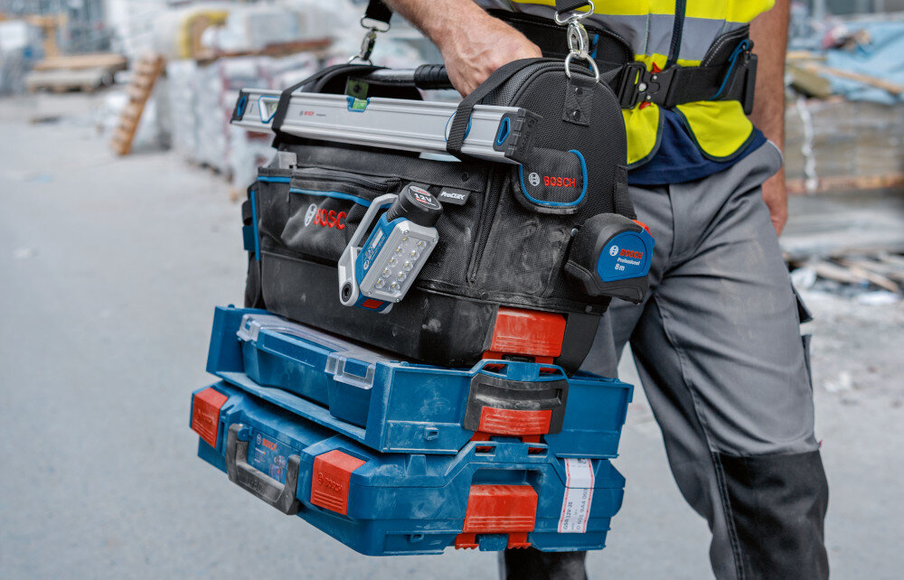 Torba narzędziowa BOSCH Professional GWT 20 plac budowy blok osoba idzie drogą na ramieniu niesie torbę wypełnioną narzędziami torba przymocowana do paska liczne przegródki i wnęki indywidualne rozmieszczenie narzędzi zintegrowane taśmy mocowanie uchwytu ProClick możliwość przypięcia torby do dowolnego paska o maksymalnej średnicy 50 mm