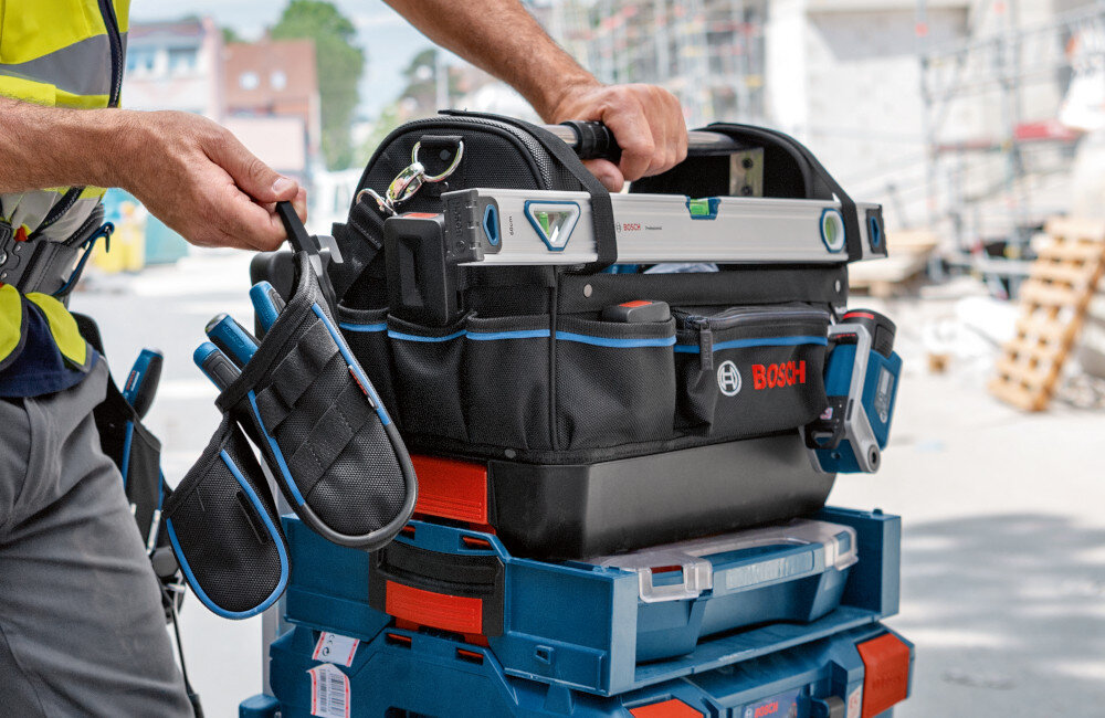 Torba narzędziowa BOSCH Professional GWT 20 plac budowy torba stoi na stosie skrzynek obok stoi osoba zintegrowane zatrzaski możliwość łączenia z walizkami L-BOXX zabezpieczenie narzędzi przed przypadkowym wypadnięciem czy uszkodzeniem