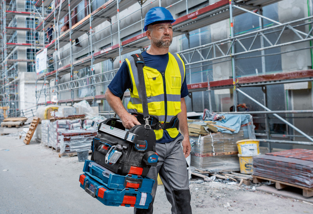 Torba narzędziowa BOSCH Professional GWT 20 plac budowy blok osoba idzie drogą na ramieniu niesie torbę wypełnioną narzędziami ergonomiczny uchwyt regulowany pas na ramię z miękką wyściółką obciążenie do 23 kg