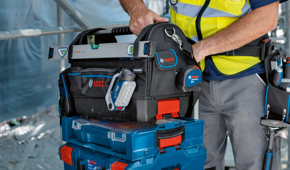 Torba narzędziowa BOSCH Professional GWT 20 plac budowy torba ustawiona na skrzynkach narzędziowych wypełniona narzędziami obok stoi osoba wymiary 230 × 430 × 340 mm waga 2,4 kg do pracy stacjonarnej i mobilnej wysoka odporność na intensywne użytkowanie i trudne warunki dla profesjonalistów i majsterkowiczów
