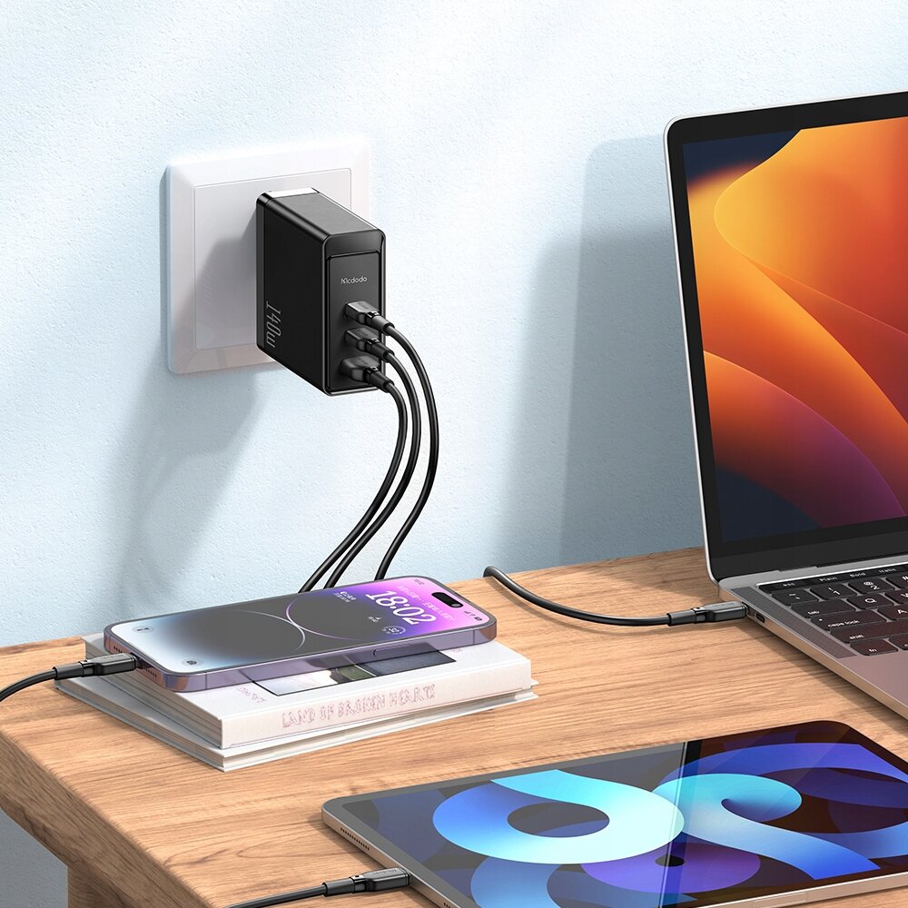 Na zdjęciu widać czarną ładowarkę sieciową Mcdodo podłączoną do gniazdka, z trzema kablami USB-C podłączonymi do laptopa, telefonu i tabletu. Na ładowarce widoczny jest napis \'Mcdodo\' oraz \'140W\'.
